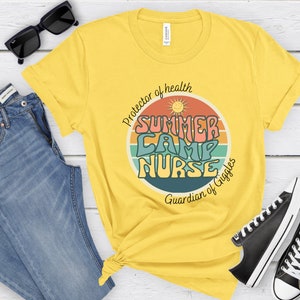 May include: Yellow t-shirt with a retro-style graphic that says "Summer Camp Nurse" in colorful letters. The graphic also includes a sun and a rainbow. The text below the graphic says "Protector of health Guardian of Giggles".