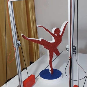 May include: A foam dancer cutout with a red glitter finish, mounted on a circular blue glitter base, positioned within a silver metal frame structure featuring red rectangular components, black cables, and threaded rods for mechanical movement.