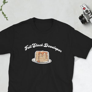 Puede incluir: Camiseta negra con un gráfico blanco de una pila de panqueques con jarabe y el texto "Full Stack Developer" en cursiva blanca.