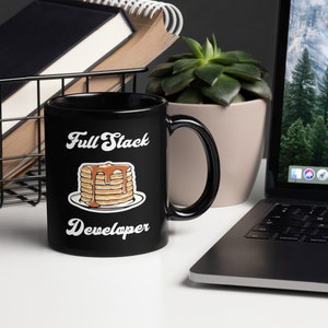 Puede incluir: Taza de café negra con un gráfico blanco de una pila de panqueques con jarabe y el texto "Full Stack Developer".