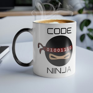 Pode incluir: Caneca de café de cerâmica branca com alça preta. A caneca tem um design de ninja preto e vermelho com o texto "CODE NINJA" e "01001110" nela.