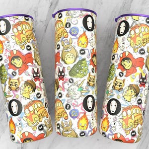 May include: Three white insulated tumblers with purple lids, decorated with colourful cartoon characters. The design includes images of a cat-like creature, a girl in a red cloak, and other whimsical figures. The tumblers are cylindrical.