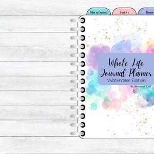 May include: A spiral-bound journal with a watercolor design. The cover features the text "Whole Life Journal Planner" in a light blue box, with colorful watercolor splashes. Tabs for months and sections are visible.