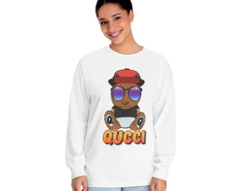 May include: White long-sleeve t-shirt with a cartoon brown bear wearing a red baseball cap and sunglasses. The bear is sitting down and the text "QUCCI" is printed below it.