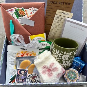 May include: A gift box filled with self-care and reading essentials. Includes a brown envelope with stickers, a teal pen, tea bags, a green floral mug, eye masks, a fuzzy sock, and a book review card. A perfect gift for book lovers.