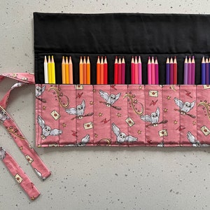 May include: A pink fabric pencil roll with a black flap and a white owl print. The roll has a tie closure and is filled with coloured pencils.