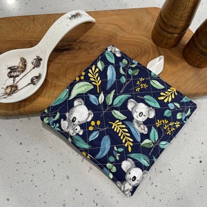 May include: A blue quilted pot holder with a pattern of koalas and eucalyptus leaves. The pot holder has a white loop for hanging.