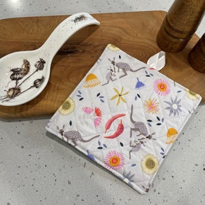 May include: A square pot holder with a quilted design. The pot holder features a white background with a colourful pattern of kangaroos, flowers, and chilli peppers.