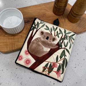 May include: A white pot holder with a black border features a cartoon koala bear sitting in a eucalyptus tree with pink flowers.