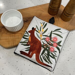 May include: A white pot holder with a kangaroo and pink flowers on a white background. The pot holder has a black trim.
