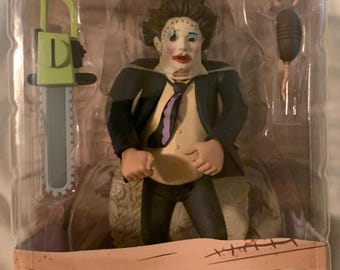 May include: A Leatherface action figure from the Texas Chainsaw Massacre film, packaged in a clear plastic case. The figure is wearing a suit and tie, with a chainsaw and a mallet accessory. The packaging includes the text "Leatherface (Pretty Woman)".