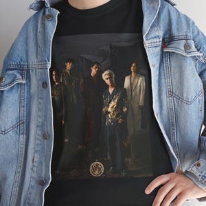 May include: Black t-shirt featuring a group of people in various outfits. The image on the shirt has a dark, moody aesthetic. The people are standing in a line, with a circular design at the bottom of the image.