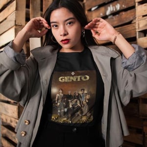 May include: A woman wearing a gray trench coat over a black t-shirt with a band graphic that says "GENTO".