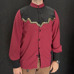 May include: A burgundy long-sleeve shirt with a black yoke and cuffs. The yoke features a gold and green geometric design. The shirt has a mandarin collar and black buttons. The sleeves are cuffed at the wrists.