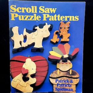 May include: A blue book cover with the title "Scroll Saw Puzzle Patterns" in yellow. The cover features wooden puzzle pieces of a cow, tiger, apple, and Native American figure. The author's names are also visible.