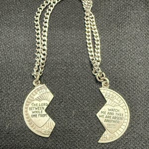 May include: A silver-toned necklace featuring two half-moon pendants that fit together. Each pendant has a chain and engraved text. One says "THE LORD BETWEEN WHILE ONE FROM" and the other says "WATCH ME AND THEE WE ARE ABSENT ANOTHER".