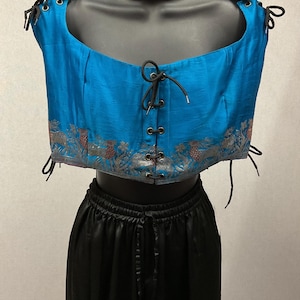 May include: A bright blue sleeveless bodice featuring a front lace-up closure with black cords, silver metal grommets, and a decorative silver and red patterned border along the bottom hem. The bodice is paired with a solid black skirt with a drawstring waist.