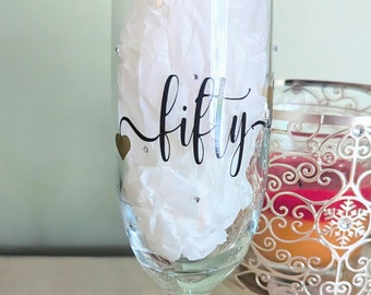 May include: A clear glass champagne flute with a black and gold design that says "fifty" on the side. The flute is decorated with black and white beads around the stem.