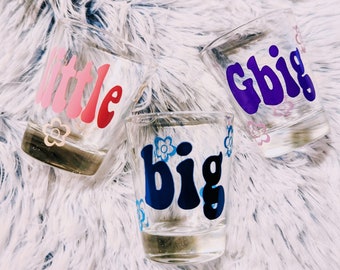 May include: Three clear glass cups with the words "little", "big", and "Gbig" written in colorful, retro-style lettering. Each cup has a small flower design.