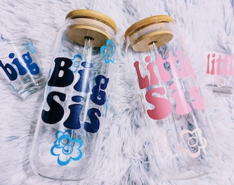 May include: Two glass water bottles with bamboo lids and straws. One bottle has a blue and white design with the text "Big Sis" and the other bottle has a pink and white design with the text "Little Sis".