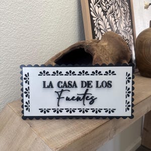 May include: A rectangular white sign with black scalloped edges and floral accents. The sign reads "LA CASA DE LOS Fuentes" in black script. The sign is displayed on a wooden surface.