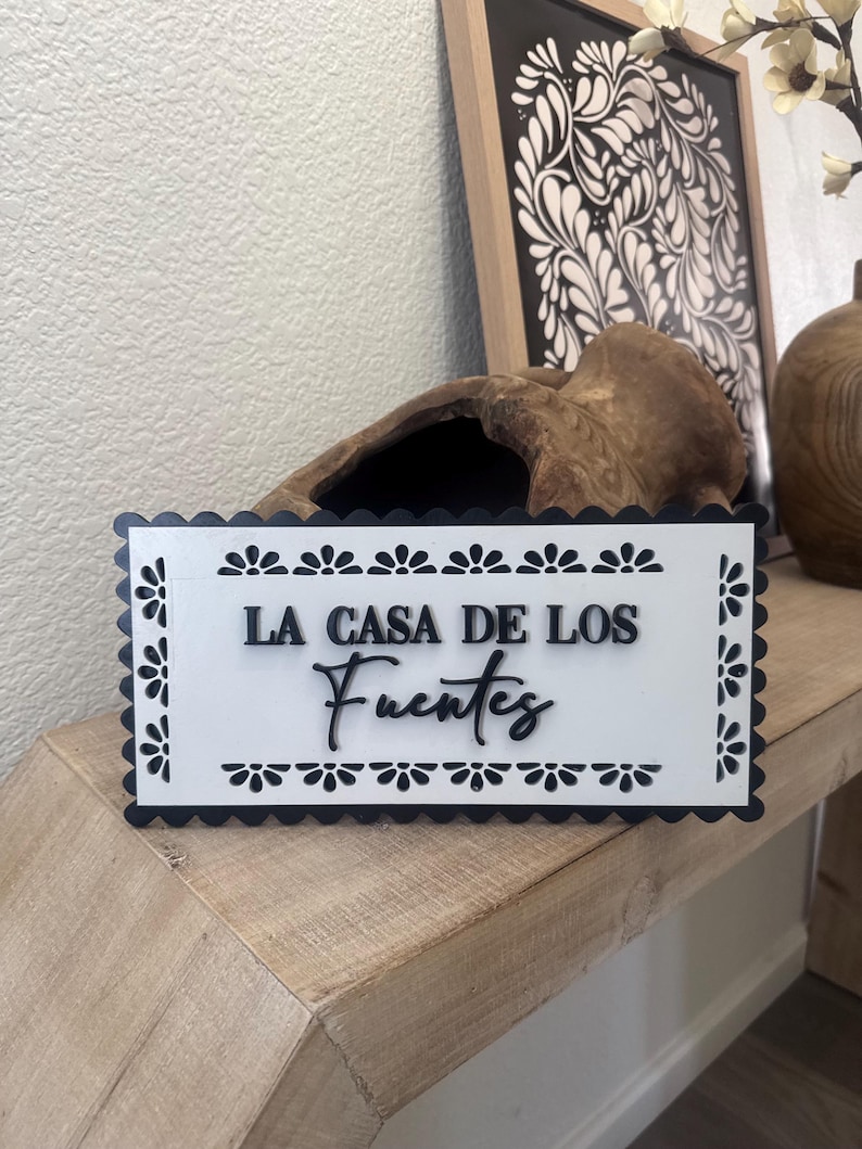 May include: A rectangular white sign with black scalloped edges and floral accents. The sign reads "LA CASA DE LOS Fuentes" in black script. The sign is displayed on a wooden surface.