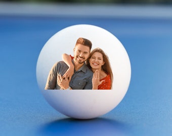 May include: A white ping pong ball with a photo of a couple smiling and hugging on it. The couple is standing in front of a blurred background.