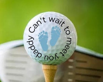 May include: A white golf ball with a blue baby footprint design and the text "Can't wait to meet you Daddy" on a golf tee.