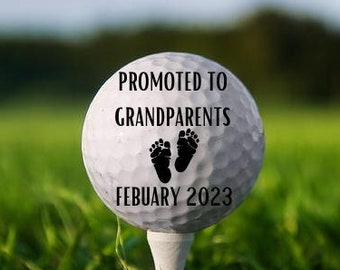 May include: A white golf ball with the text "Promoted to Grandparents February 2023" and two black footprints on a green grassy field.