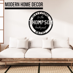 May include: Black metal sign with the text "Welcome to the Thompson backyard patio" in a circle. The sign is hanging on a white wall above a white couch.