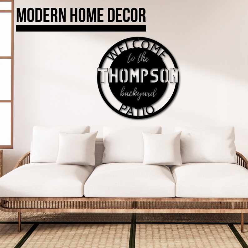 May include: Black metal sign with the text "Welcome to the Thompson backyard patio" in a circle. The sign is hanging on a white wall above a white couch.