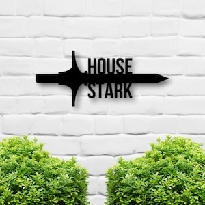 May include: Black metal wall decor featuring a sword with the words "House Stark" on a white brick wall background.