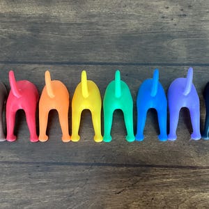 May include: A row of colorful dog butt figurines in shades of brown, red, orange, yellow, green, blue, purple, and black. Each figurine has a tail and is positioned on a wooden surface.