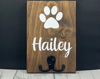 May include: A brown wooden dog leash hook with a white paw print and the name "Hailey" written on it.