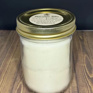 May include: A clear glass jar candle with a gold lid. The candle is filled with a white wax and has a label that reads "Paarsitive Vibes." The label also states the candle is hand-poured in NJ and has a net weight of 16 oz.