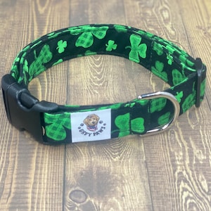 May include: A black dog collar with a green shamrock pattern. The collar has a black plastic buckle and a silver metal D-ring. A white label with a dog graphic and the text "GOOFY PAWS" is attached to the collar.