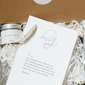 May include: A gift box containing a jar of calendula salve with a silver lid, a dried wheat stalk, and a white card featuring a line drawing of a person holding a baby and a printed note from Hannah to Jay about motherhood and relaxation.