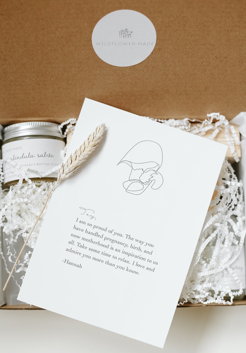 May include: A gift box containing a jar of calendula salve with a silver lid, a dried wheat stalk, and a white card featuring a line drawing of a person holding a baby and a printed note from Hannah to Jay about motherhood and relaxation.