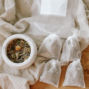 May include: A white marble mortar filled with a blend of dried green herbs and yellow flower petals, accompanied by four small, translucent white drawstring tea bags and a clear plastic pouch containing a white label with printed text.