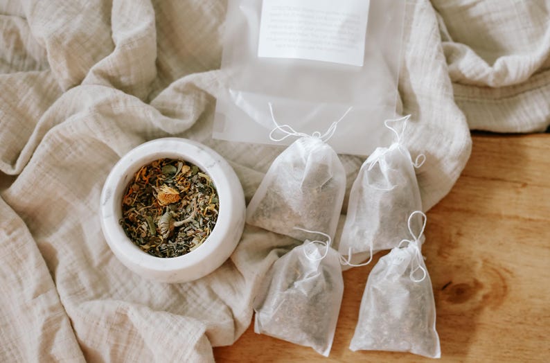 May include: A white marble mortar filled with a blend of dried green herbs and yellow flower petals, accompanied by four small, translucent white drawstring tea bags and a clear plastic pouch containing a white label with printed text.