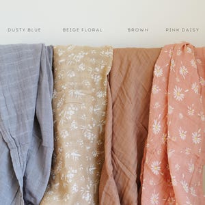 Peut inclure: Quatre langes en mousseline pli&eacute;s c&ocirc;te &agrave; c&ocirc;te. Les couleurs et motifs sont &eacute;tiquet&eacute;s Dusty Blue, Beige Floral avec des fleurs blanches, Brown uni, et Pink Daisy avec un imprim&eacute; floral blanc et jaune sur un fond rose.