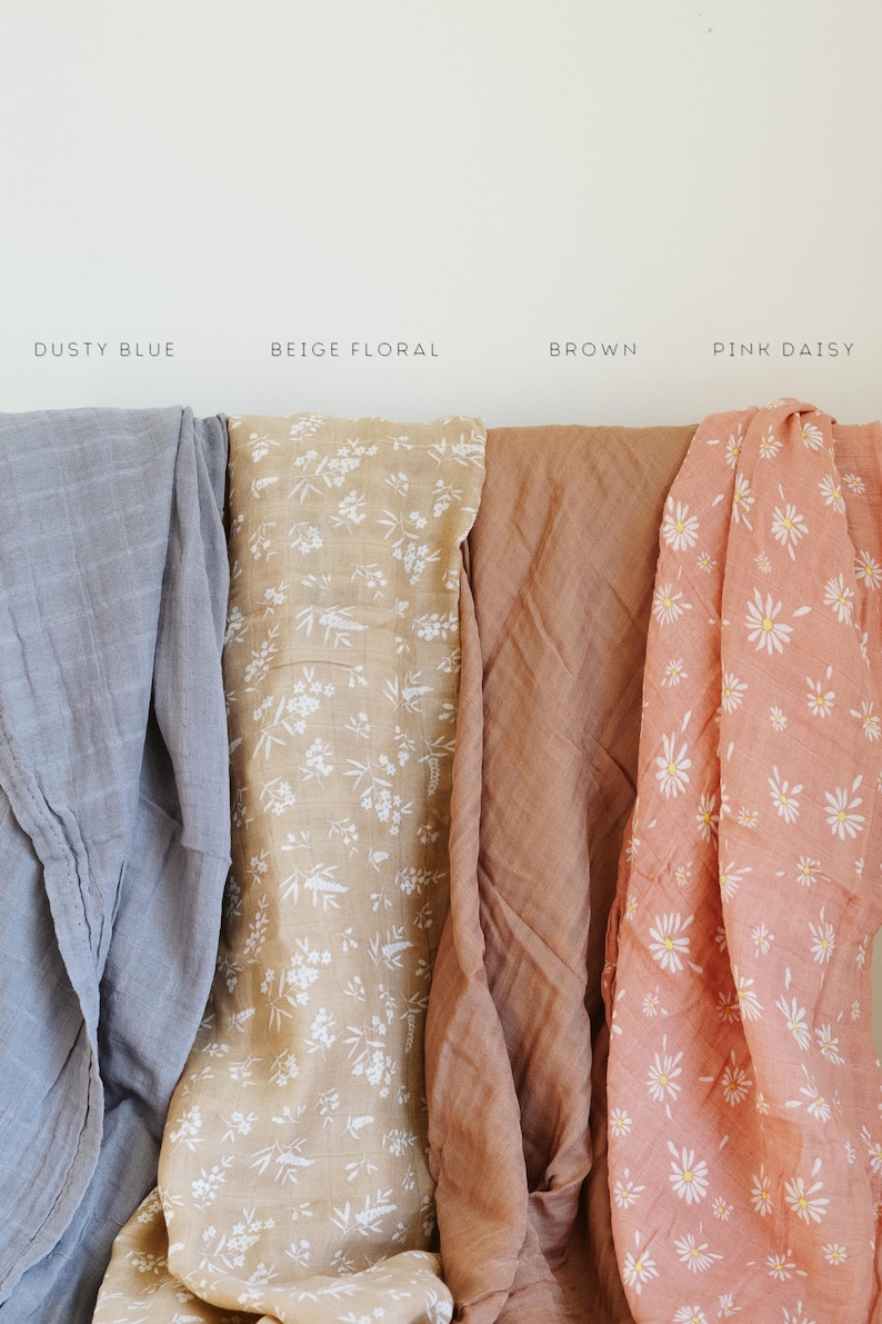 Peut inclure: Quatre langes en mousseline pli&eacute;s c&ocirc;te &agrave; c&ocirc;te. Les couleurs et motifs sont &eacute;tiquet&eacute;s Dusty Blue, Beige Floral avec des fleurs blanches, Brown uni, et Pink Daisy avec un imprim&eacute; floral blanc et jaune sur un fond rose.