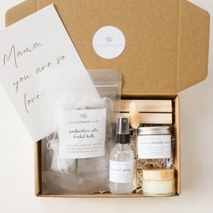 May include: A brown cardboard gift box containing a clear pouch of postpartum sitz herbal bath, a spray bottle, a small jar of calendula salve, a small cream jar, and a white card with the text Mama, you are so loved, plus a dried floral stem.