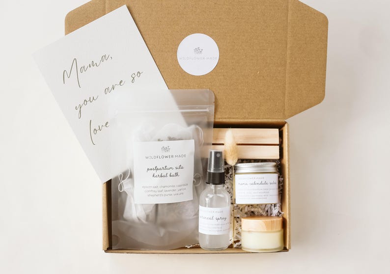 May include: A brown cardboard gift box containing a clear pouch of postpartum sitz herbal bath, a spray bottle, a small jar of calendula salve, a small cream jar, and a white card with the text Mama, you are so loved, plus a dried floral stem.