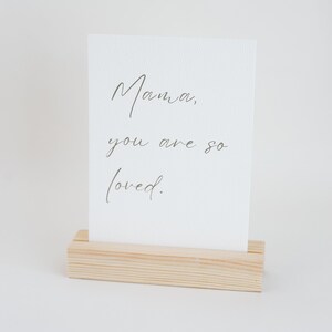 May include: A rectangular white card with a textured surface featuring the dark grey cursive text Mama, you are so loved. The card is held upright by a light-colored rectangular wooden block stand with a horizontal slot cut into the top center.
