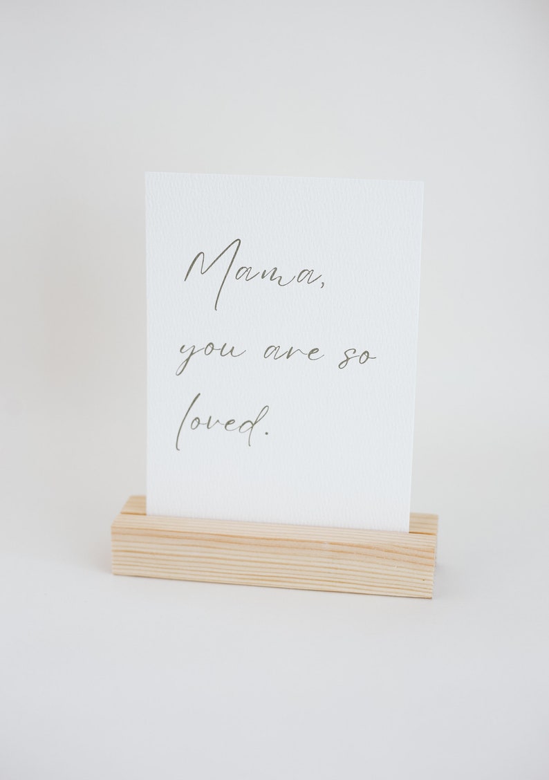 May include: A rectangular white card with a textured surface featuring the dark grey cursive text Mama, you are so loved. The card is held upright by a light-colored rectangular wooden block stand with a horizontal slot cut into the top center.