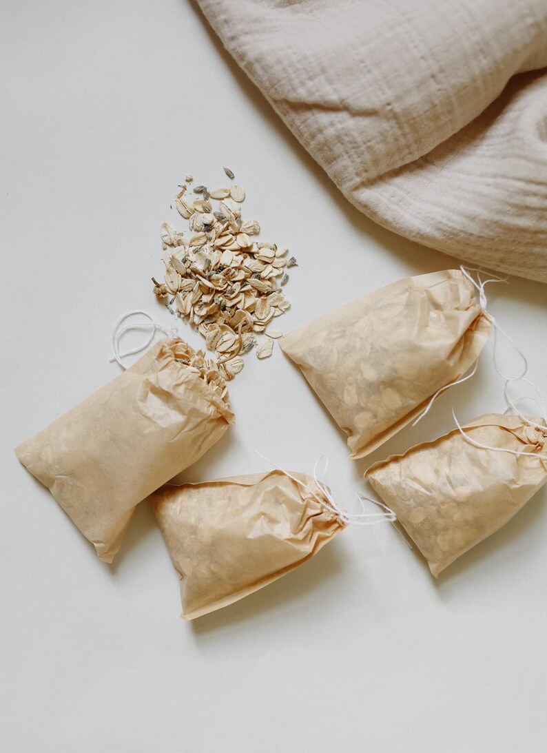 Peut inclure: Quatre petits sachets rectangulaires en papier brun clair avec des fermetures &agrave; cordon blanc, remplis d'avoine s&eacute;ch&eacute;e et de boutons de lavande. Un tas de ce m&ecirc;me m&eacute;lange d'avoine et de lavande est dispos&eacute; &agrave; c&ocirc;t&eacute;, avec un tissu beige.