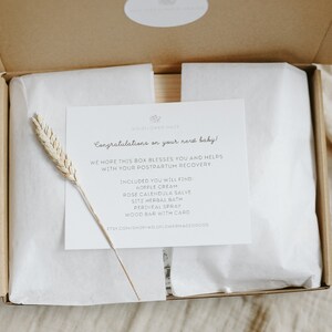 May include: A cardboard gift box containing white tissue paper and a white card with black text that reads: Congratulations on your new baby! We hope this box blesses you and helps with your postpartum recovery. Included: nipple cream, rose calendula salve, sitz herbal bath, perineal spray, wood bar with card. A dried wheat stalk rests on the card.