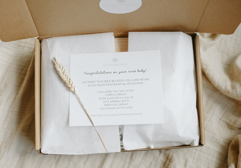 May include: A cardboard gift box containing white tissue paper and a white card with black text that reads: Congratulations on your new baby! We hope this box blesses you and helps with your postpartum recovery. Included: nipple cream, rose calendula salve, sitz herbal bath, perineal spray, wood bar with card. A dried wheat stalk rests on the card.