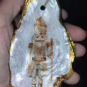 May include: A Christmas ornament featuring a painted nutcracker soldier on a white surface, set within a gold-edged oyster shell. The ornament has a small hole for hanging.